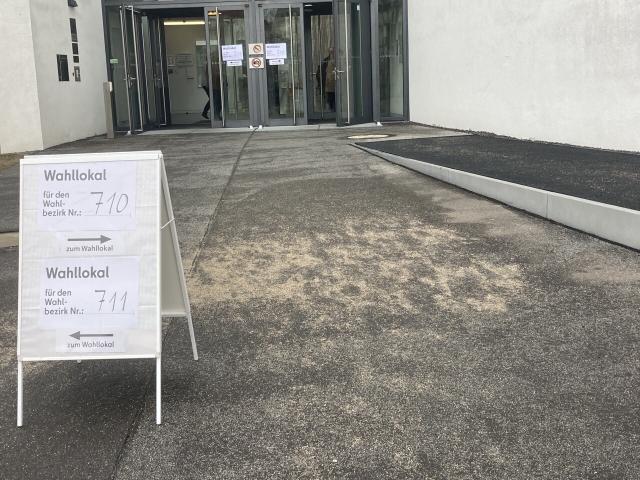 A polling station at a school in Germany. We see the front entrance in the distance. More proximally, a small poster stand informing voters where to go. No people standing in line. 
