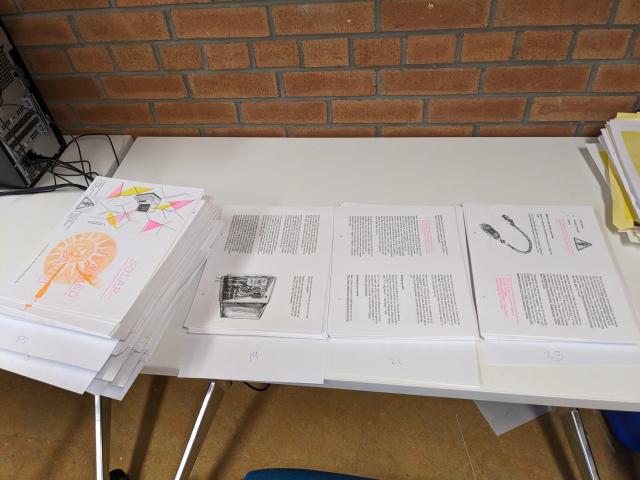 A table with piles of paper, separated out by page numbers. The title says Solar-Powered Media and has bright pink text and images of a sun with an Ethernet cable for rays. The rest of black with grayscale illustrations of tech diagrams and pink accent text