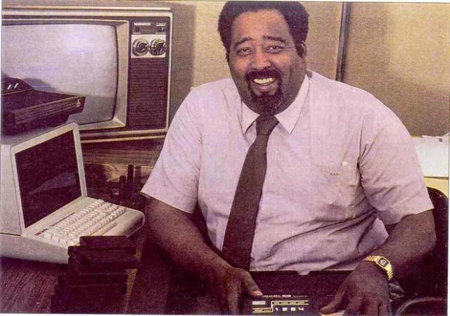 Console inventor Jerry Lawson sits at desk with computer, smiling, holding a video game console