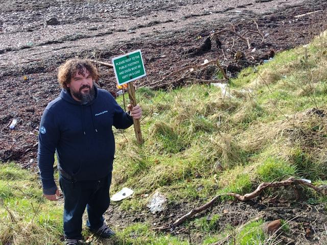 Me holding a sign to stay on path on a plastic-covered pebble beach