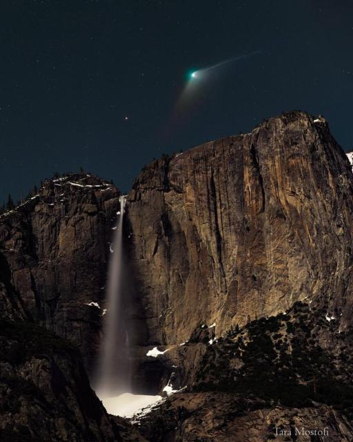 A comet with a green head and extended tails is seen above a high water fall. In the night sky field just above the falls, an orange dot -- the star Kochab -- is visible.