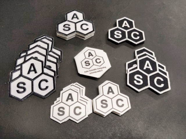 Stickers and patches with the ASC hexagonal logo laid out on a table.  All stickers and some patches have a white background and black letters.  Some patches have a black background and white letters.